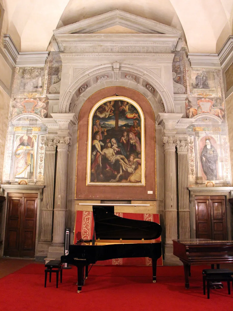 The stone-framed altar wall of Santa Monaca church with a painted altarpiece of the Lamentation in an arched frame, painted saints in niches either side and a grand piano below