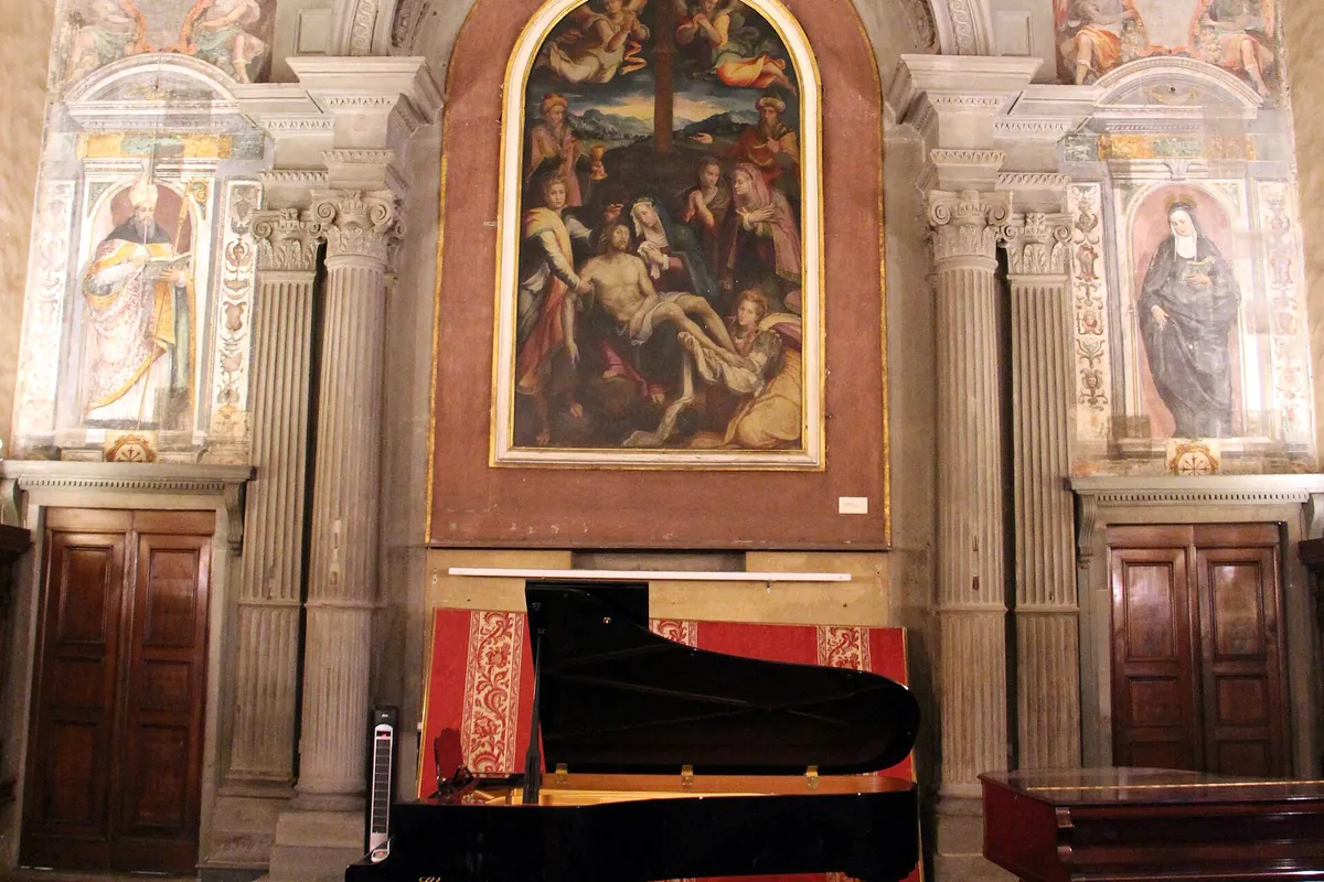 The stone-framed altar wall of Santa Monaca church with a painted altarpiece of the Lamentation in an arched frame, painted saints in niches either side and a grand piano below