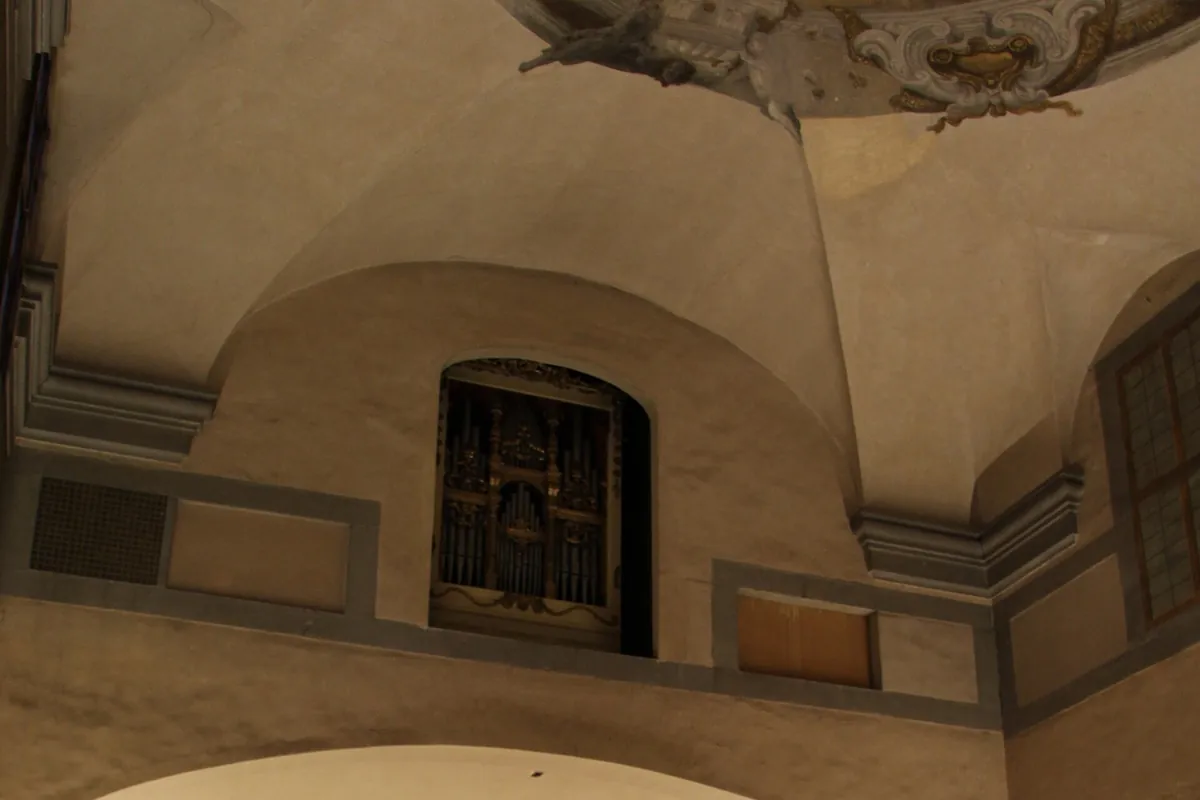 A window high on the wall of the nave of Santa Monaca church holding an organ, beneath a vaulted ceiling with grey stone mouldings