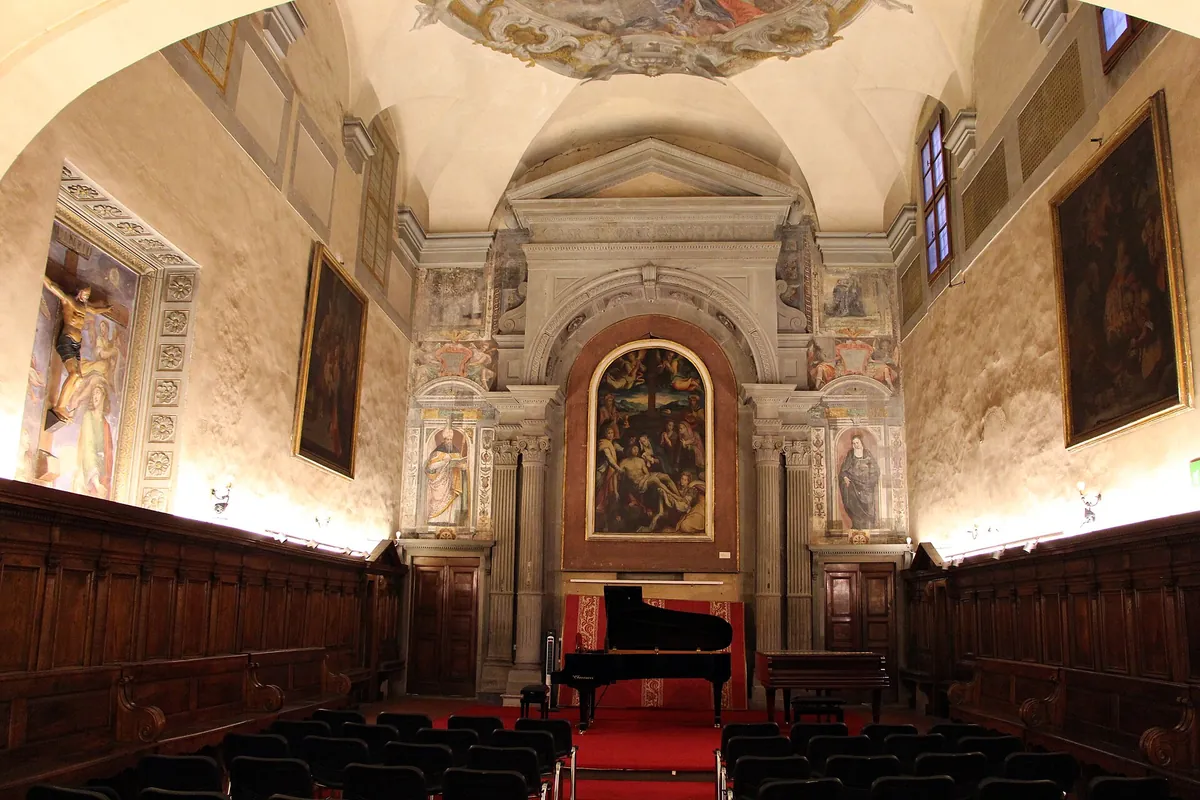 The nave of Santa Monaca church set for a concert, with rows of chairs facing a grand piano on a red carpet in front of a stone-framed altar and wooden choir stalls along both walls