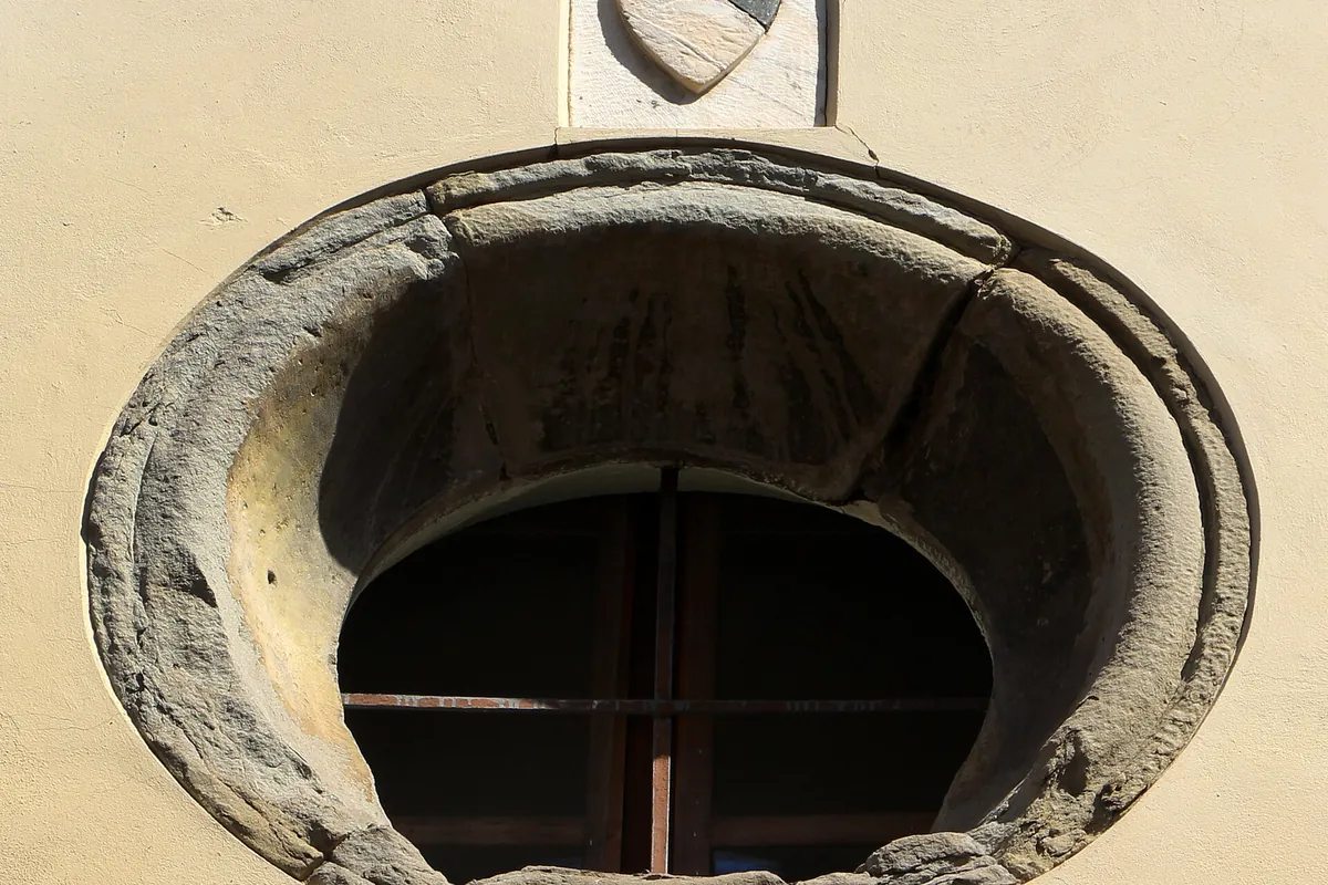A round stone window set in a pale plastered wall on Via Santa Monaca in Florence, with a small stone shield above it